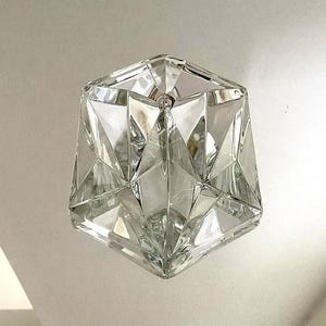 Op de afbeelding: Een heldere, facetgeslepen glazen lamp met een geometrisch ontwerp. De lamp heeft een zeshoekige vorm met meerdere driehoekige facetten, waardoor een sprankelend effect ontstaat. De lichtbron is van binnen zichtbaar.