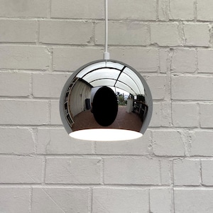 Puede incluir: Una lámpara colgante esférica cromada cuelga de un cable blanco contra una pared de ladrillos blancos. La superficie reflectante de la lámpara refleja el entorno. La luz emite un brillo suave desde la parte inferior. Diseño moderno.