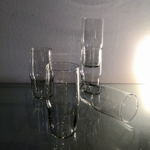 May include: Five clear glass drinking glasses of various sizes and shapes are stacked and arranged on a reflective surface.
