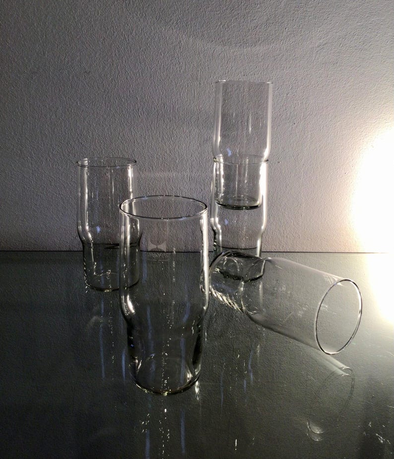 May include: Five clear glass drinking glasses of various sizes and shapes are stacked and arranged on a reflective surface.