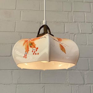May include: A vintage pendant light fixture with a white, rounded shade. The shade features a hand-painted design of orange and red leaves and berries. The fixture is suspended by a white cord and has a brass-colored metal bracket.