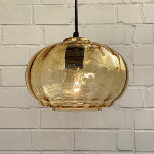 Könnte beinhalten: Eine Hängeleuchte mit einem kugelförmigen, bernsteinfarbenen Glasschirm. Der Schirm hat ein strukturiertes, wellenförmiges Muster und ist an einem schwarzen Kabel aufgehängt. Die Glühbirne ist im Inneren des Schirms sichtbar und beleuchtet die Leuchte.