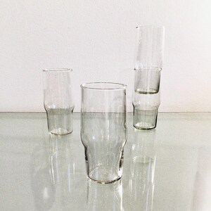 May include: Four clear glass drinking glasses, three stacked on top of each other. The glasses are tall and cylindrical with a slightly flared lip.