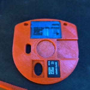 May include: Orange 3D printed case with two slots for memory cards. One slot is for a Micro Center 4GB Micro SD card and the other slot is for a Micro SD card with the text "Memory Stick Pro Duo" printed on it.