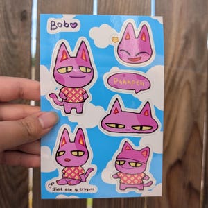 May include: A sticker sheet featuring Bob, a pink cat character, with various poses and expressions. The sheet includes the text "Bob" and "Pehhpeh". The cat is wearing a patterned shirt in some images. The text "Just ate 4 crayons" is also included.