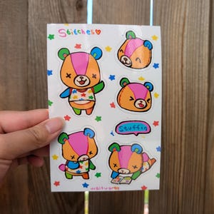 May include: A sticker sheet featuring cartoon bear characters in various poses. The bears have orange, pink, and blue color schemes. The text "Stitches" and "Stuffin" are included. The stickers are decorated with stars and are held against a wooden background.