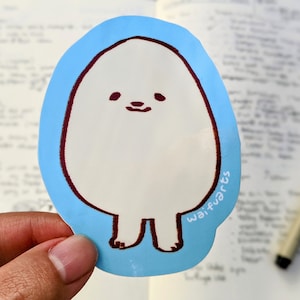 Op de afbeelding: Een gestanste sticker met een cartoonachtig, eivormig personage met een lachend gezicht en kleine voetjes. Het personage is wit met een bruine omtrek en details, tegen een lichtblauwe achtergrond. De sticker heeft de tekst "waifvarts" in het wit.