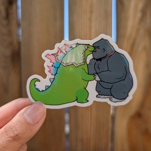 Puede incluir: Un dinosaurio verde con un velo blanco y rosa besa a un gorila gris con una pajarita negra. El dinosaurio y el gorila son personajes de dibujos animados.