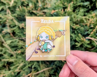 Op de afbeelding: Een gouden emaille pin met een cartoonachtige afbeelding van Zelda uit de Legend of Zelda-videogameserie. Zelda draagt een groen-witte jurk en houdt een klein geel vogeltje vast. De pin is op een witte kaart met de tekst "ZELDA" en "by waifuarts".