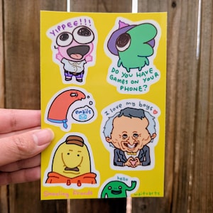 May include: A yellow sticker sheet featuring cartoon characters with various expressions and text. The text includes "YIPPEE!!!", "emails", "Do you have games on your phone?", "I love my boys", and "hello". The sticker sheet is held by a hand.