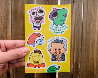 Pode incluir: Uma folha de adesivos amarela com personagens de desenhos animados e texto. O texto inclui "YIPPEE!!!", "emails", "Do you have games on your phone?", "I love my boys" e "hello". A folha de adesivos é segurada por uma mão.