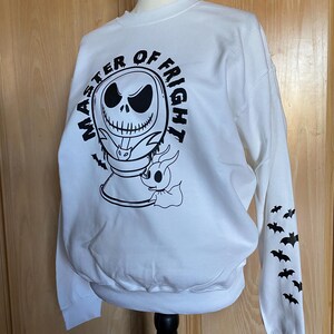 May include: White sweatshirt with a black and white graphic of Jack Skellington from The Nightmare Before Christmas. The graphic says "Master of Fright" and features a small ghost. The sleeves have a repeating pattern of black bats.