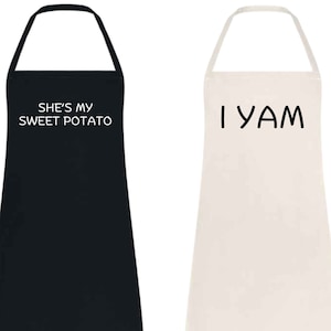 May include: Two aprons, one black and one off-white. The black apron has the text "SHE'S MY SWEET POTATO" in white. The off-white apron has the text "I YAM" in black. Both aprons have a halter neck and a long, rectangular body.