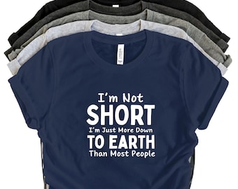 Puede incluir: Una pila de camisetas en varios colores, incluyendo azul marino, negro y gris. La camiseta azul marino de enfrente tiene el texto blanco "I'm Not SHORT I'm Just More Down TO EARTH Than Most People."