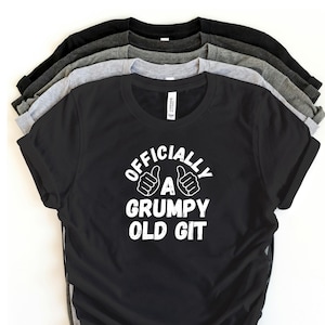 Può includere: Una pila di magliette in varie tonalità di grigio e nero, con una maglietta nera davanti. La maglietta anteriore ha il testo bianco "OFFICIALLY A GRUMPY OLD GIT", con grafiche di pollice in su.