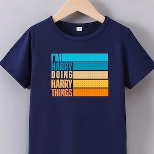Puede incluir: Camiseta azul marino de manga corta con el texto "I'M HARRY DOING HARRY THINGS" en un diseño a rayas colorido. La camiseta está colgada en una percha de madera.