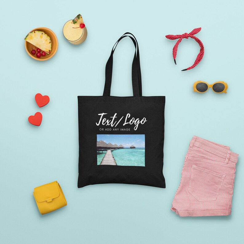 K&ouml;nnte beinhalten: Schwarze Tragetasche mit wei&szlig;em Text, der "Text/Logo or add any image" lautet. Die Tasche hat ein tropisches Strandmotiv darauf gedruckt. Die Tasche ist von verschiedenen Gegenst&auml;nden umgeben, darunter ein Ananas, ein Getr&auml;nk, ein Stirnband, eine Sonnenbrille, Herzen und eine kleine Handtasche.
