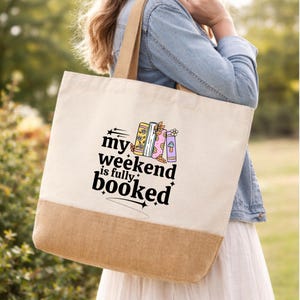 May include: A beige canvas tote bag with a brown base and handles. The bag displays the text "my weekend is fully booked" in black, accompanied by colourful illustrations of books and a mushroom.