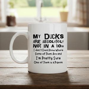 Könnte beinhalten: Weiße Keramiktasse mit gebogenem Henkel. Die Tasse hat einen schwarzen Text mit der Aufschrift "MY DUCKS ARE ABSOLUTELY NOT IN A ROW". Zusätzlicher Text lautet "I don't Even Know Where Some of Them Are and I'm Pretty Sure One of Them is a Pigeon."