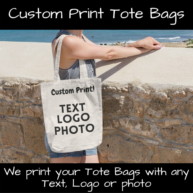 K&ouml;nnte beinhalten: Eine wei&szlig;e Canvas-Tragetasche mit schwarzem Text, der "Custom Print!"  "TEXT" "LOGO" "PHOTO" lautet. Die Tasche wird von einer Person gehalten, die in der N&auml;he einer Steinmauer und des Meeres steht.