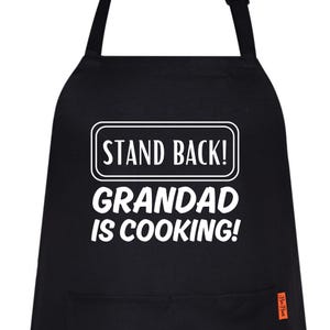 Puede incluir: Delantal negro con texto blanco que dice: "STAND BACK! GRANDAD IS COOKING!". El delantal tiene un bolsillo y una correa para el cuello. El delantal está hecho de un material duradero.