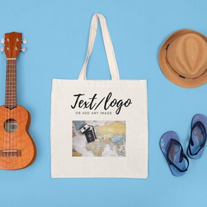 K&ouml;nnte beinhalten: Eine wei&szlig;e Canvas-Tragetasche mit dem Aufdruck "Text/logo or add any image". Die Tasche ist von verschiedenen Gegenst&auml;nden umgeben, darunter eine Ukulele, ein Strohhut und Flip-Flops.