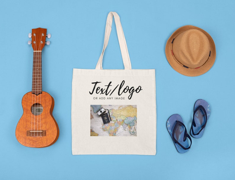 K&ouml;nnte beinhalten: Eine wei&szlig;e Canvas-Tragetasche mit dem Aufdruck "Text/logo or add any image". Die Tasche ist von verschiedenen Gegenst&auml;nden umgeben, darunter eine Ukulele, ein Strohhut und Flip-Flops.