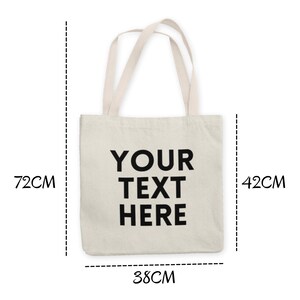K&ouml;nnte beinhalten: Eine wei&szlig;e Canvas-Tragetasche mit schwarzen Henkeln und dem aufgedruckten Text "YOUR TEXT HERE" auf der Vorderseite. Die Tasche ist 38 cm breit, 42 cm hoch und 7,5 cm tief.