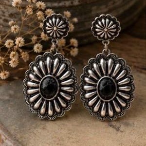 May include: A pair of silver-toned drop earrings featuring a circular top stud and a larger oval pendant. Each piece displays a sunburst design with radiating petals surrounding a smooth, black oval stone center, finished with an oxidized metal texture.