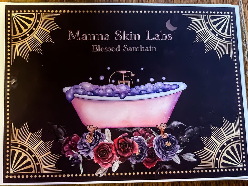 May include: A decorative card with the text "Manna Skin Labs Blessed Samhain" features a pink bathtub filled with purple bubbles. The card is framed with gold sunburst and floral accents, with a dark background.