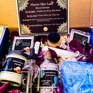 May include: A gift box from Manna Skin Labs, containing Oatmilk and Honey Body Butter, Chakra Oil, Rosehip Oil, and Abundance Milk Bath. The box includes a variety of self-care products, with a focus on natural ingredients and ritual skincare. The box is decorated with dried flower petals.