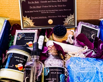 May include: A gift box from Manna Skin Labs, containing Oatmilk and Honey Body Butter, Chakra Oil, Rosehip Oil, and Abundance Milk Bath. The box includes a variety of self-care products, with a focus on natural ingredients and ritual skincare. The box is decorated with dried flower petals.