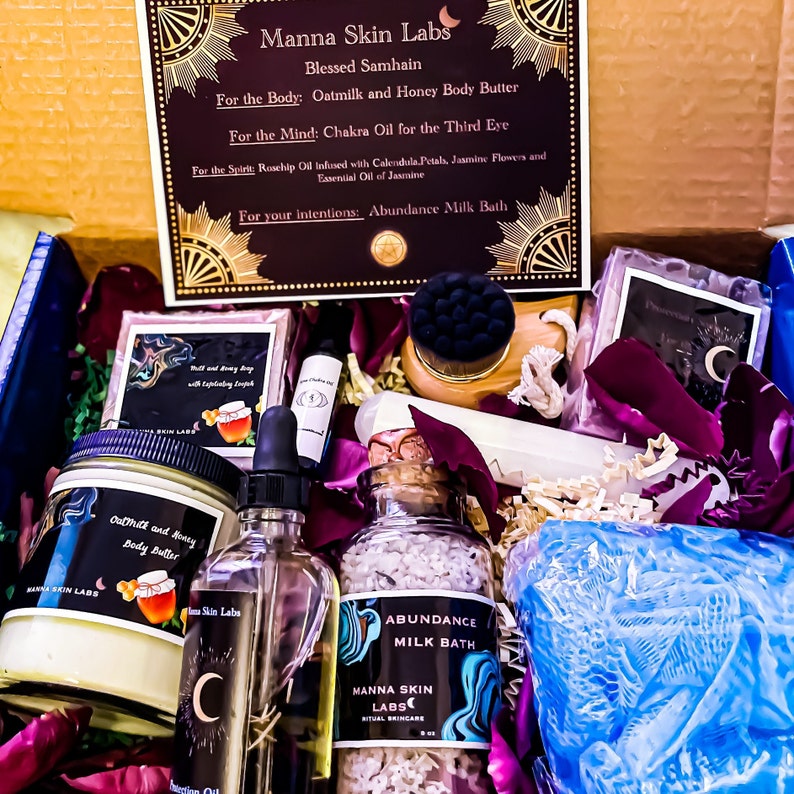 May include: A gift box from Manna Skin Labs, containing Oatmilk and Honey Body Butter, Chakra Oil, Rosehip Oil, and Abundance Milk Bath. The box includes a variety of self-care products, with a focus on natural ingredients and ritual skincare. The box is decorated with dried flower petals.