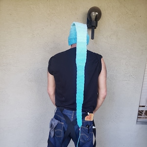 Puede incluir: Una persona lleva un gorro de punto azul y blanco con una cola larga que se extiende hasta la cintura. Lleva una camiseta negra y pantalones vaqueros azules.