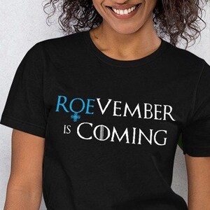 Peut inclure: T-shirt noir avec l'inscription "ROEVEMBER is COMING" en blanc et bleu. Le t-shirt est à manches courtes et a un col rond. Le texte est centré sur la poitrine.