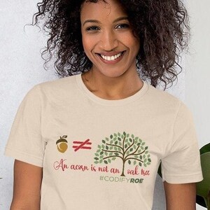 Peut inclure: T-shirt crème avec l'inscription "An acorn is not an oak tree" et un graphisme de gland et d'arbre. Le t-shirt comprend également le hashtag #CODIFYROE dans un schéma de couleurs vert et rouge.