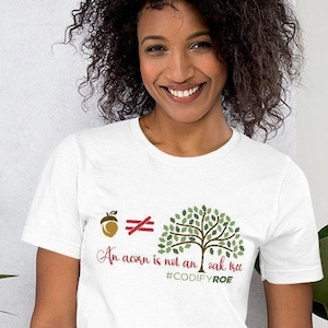 Peut inclure: T-shirt blanc avec un motif graphique représentant un gland, un arbre et le texte "An acorn is not an oak tree" en rouge et vert. Le t-shirt comprend également le hashtag #CODIFYROE.
