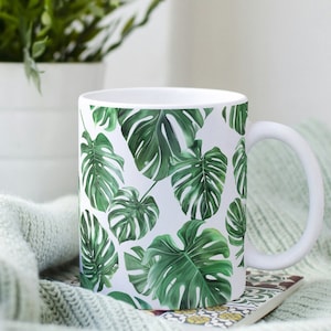 May include: White ceramic mug with a tropical leaf pattern. The leaves are green and white and are arranged in a repeating pattern.