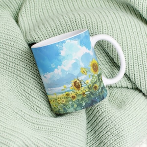 May include: White ceramic mug with a blue sky and field of sunflowers painted on it. The mug is sitting on a light green knitted blanket.