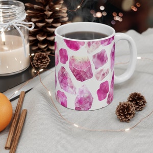 May include: White ceramic mug with a pink watercolor design of gemstones. The mug is filled with coffee.