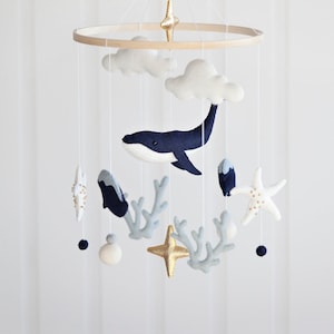 May include: A white, blue and gold baby mobile featuring a whale, clouds, starfish and coral. The mobile hangs from a wooden ring.