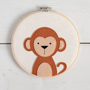 May include: A brown felt monkey with a light brown belly and black eyes is stitched onto a white felt circle. The circle is framed in a wooden hoop.