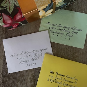 May include: An overhead shot shows three envelopes with handwritten addresses in black ink. The envelopes are light green, white, and yellow. A floral patterned box with a gold ribbon is in the background. The addresses include names, street addresses, and zip codes.