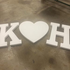 以下が含まれることがあります： 横一列に並べられた大きな白い大文字のK、白いハートの形、そして大きな白い大文字のH。これら3つの独立した装飾品は、厚みのあるブロック体フォントで、全体が滑らかで無地の白い仕上げになっています。
