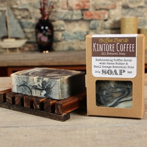 May include: A brown cardboard box with a white label that reads "Coffee Scrub KINTORRE COFFEE All Natural Soap Exfoliating Coffee Scrub with Cocoa Butter & Basil Orange Essential Oils 120g SOAP BAR". The box contains a bar of soap with a swirl pattern of black, grey, and white. The soap is sitting on a wooden soap dish.