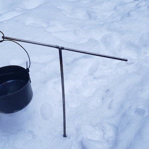 May include: A black metal pot hanging from a metal tripod stand over a snowy surface.
