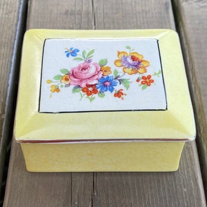 Peut inclure: Petite boîte à bijoux rectangulaire en céramique jaune. Le couvercle est orné d'un motif floral sur fond blanc, avec des roses roses, des fleurs bleues et jaunes. La boîte est bordée d'une bordure argentée.