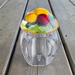 May include: A clear glass jar with a dome-shaped lid. The lid is decorated with a yellow peach, a yellow plum, and a red raspberry, all encased in resin. The jar has a textured, faceted design.