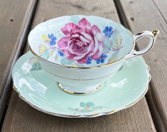 May include: A vintage teacup and saucer set. The teacup features a large pink rose design with blue and yellow accents on a light green background. The set has gold trim and a scalloped edge.
