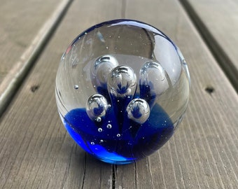May include: A clear glass paperweight with a spherical shape. The paperweight features a vibrant blue base and several silver-colored, bubble-like inclusions. The glass is smooth and reflects light, creating a decorative effect.